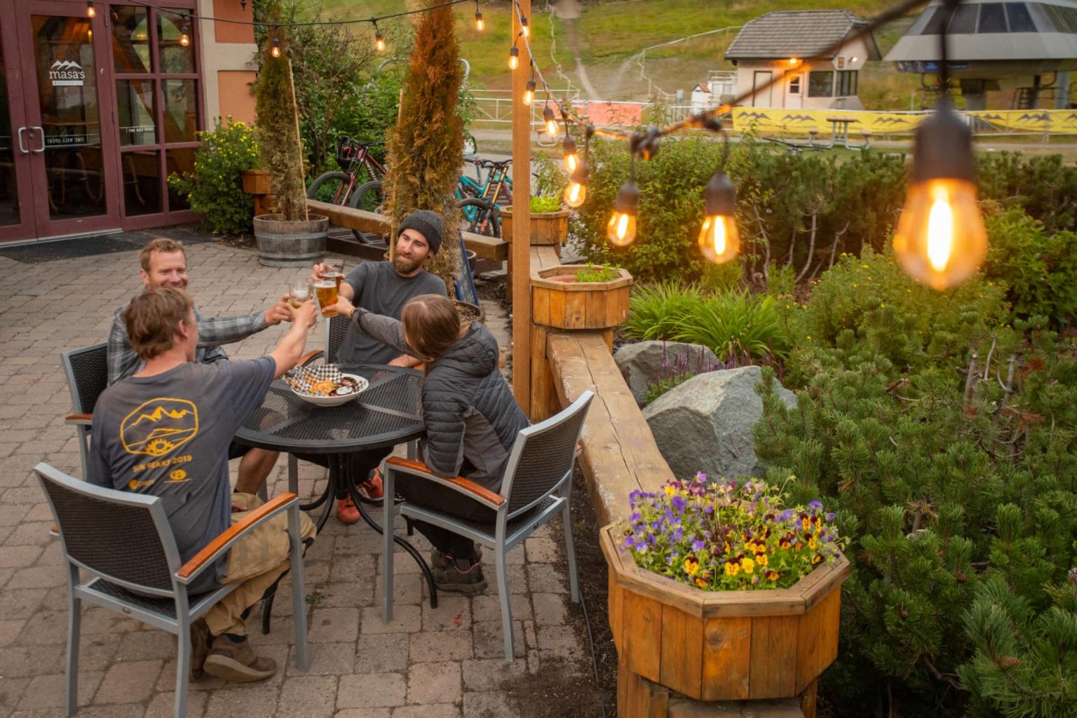 Group toasting at outdoor cafe under string lights, surrounded by greenery.