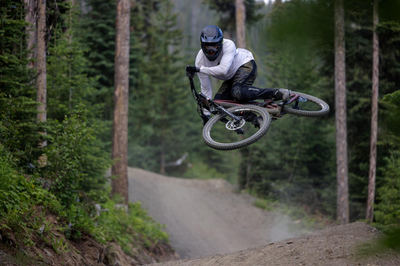 Cyclist mid-air on mountain trail, surrounded by trees.