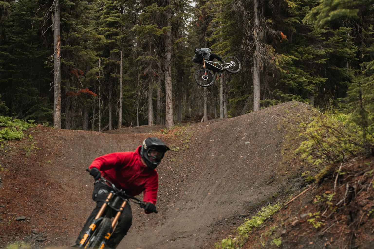 Two mountain bikers in wooded area, one in red mid-turn, another airborne.