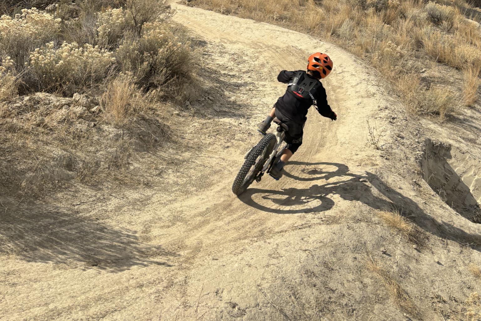 Cyclist in orange helmet rides on a dirt trail in a dry, rocky landscape.