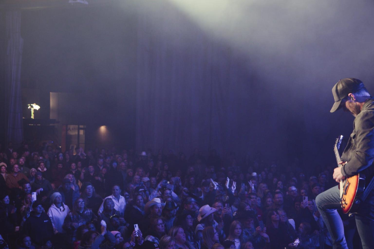 Guitarist on stage with a large crowd under blue lighting.