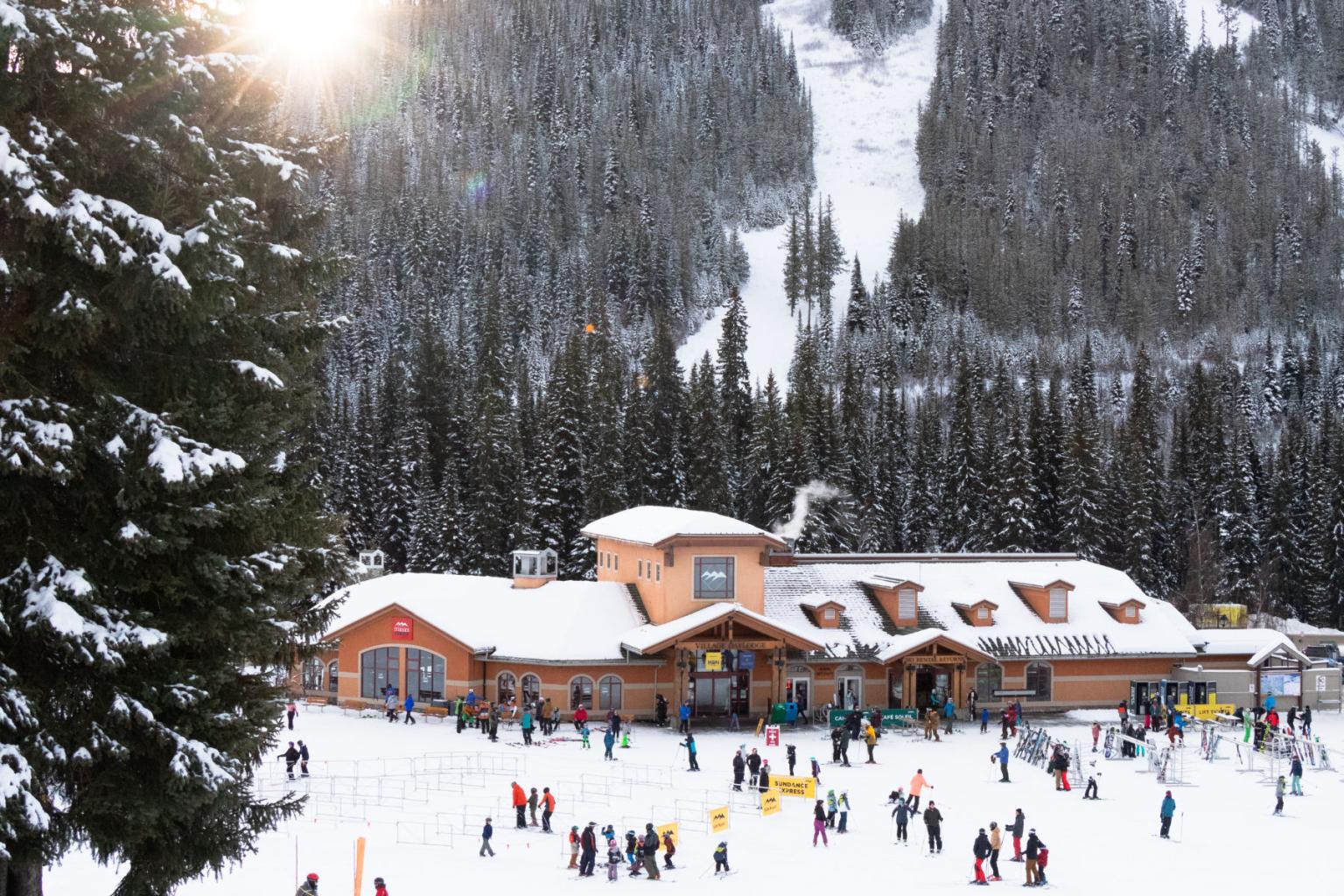 Snowy ski resort with lodge and scattered skiers; mountain and sun in background.
