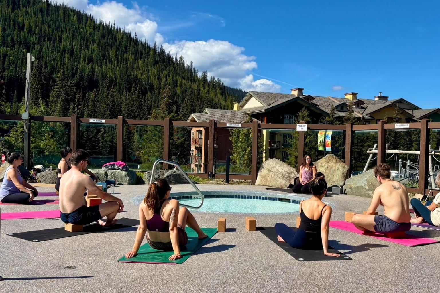 People doing outdoor yoga near a hot tub, mountains and blue sky in the background.