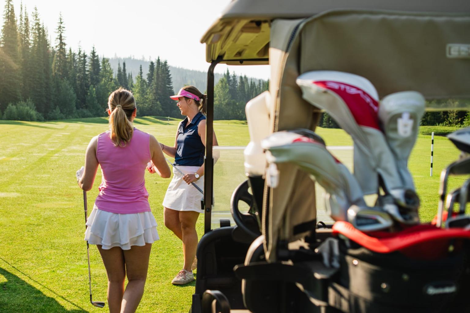 Two women talking beside a golf cart on a sunny course.