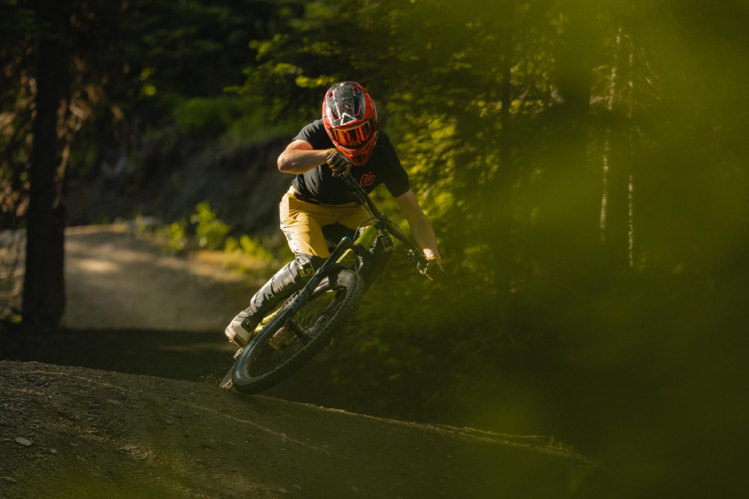 Mountain biker jumping off a dirt trail in the forest, wearing a red helmet.