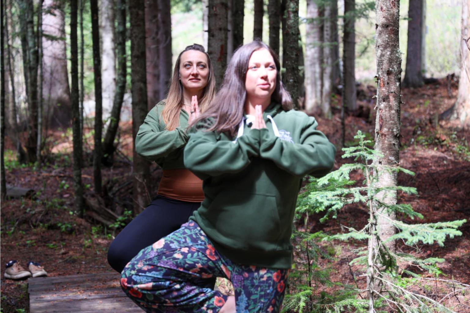 Two women practicing yoga poses in a sunlit forest.