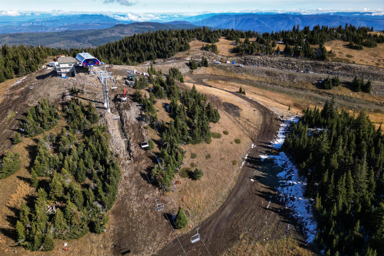 Mountain construction site with trees, machinery, and distant hills.
