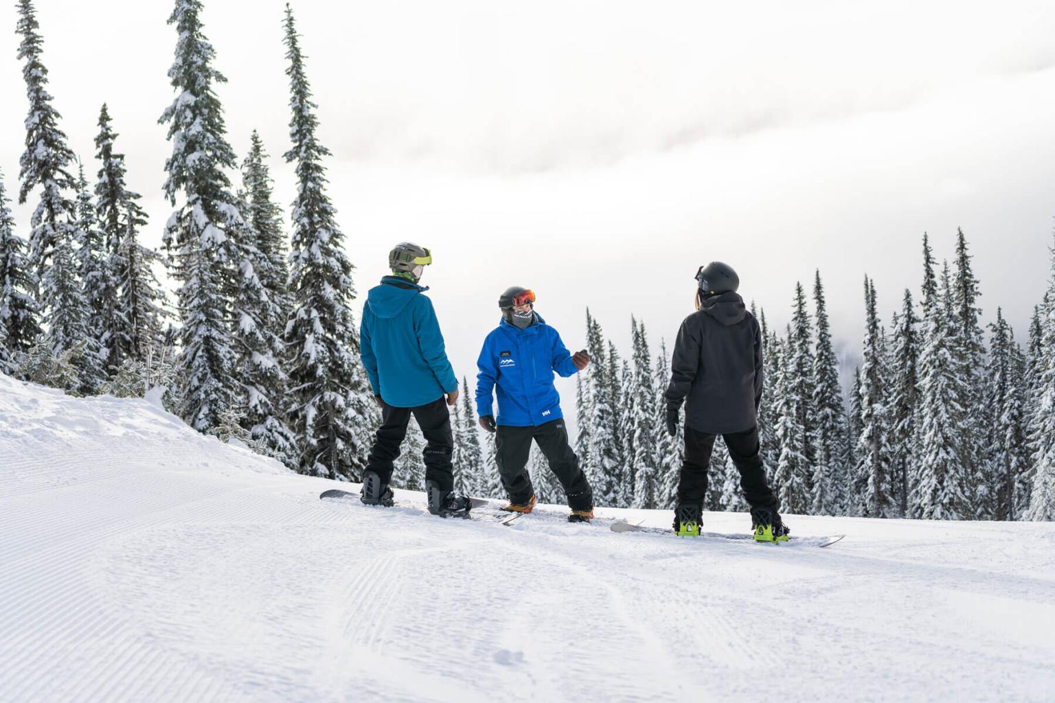 Three snowboarders chatting on a snowy slope with trees in the background.