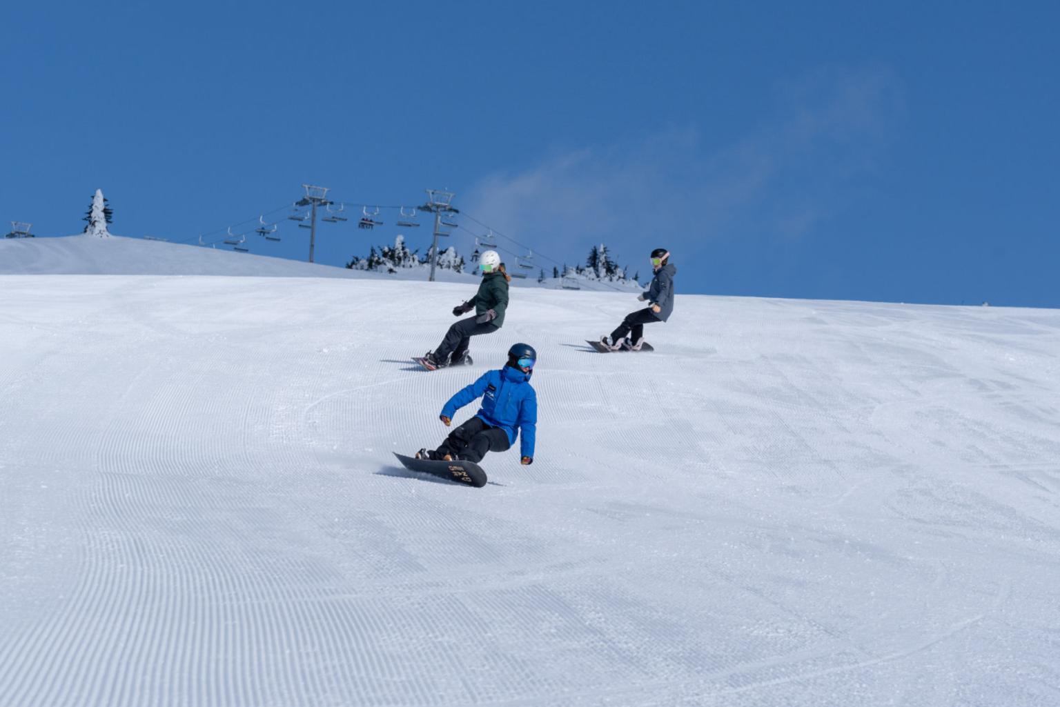 Snowboarders descending a snowy slope under a clear blue sky.
