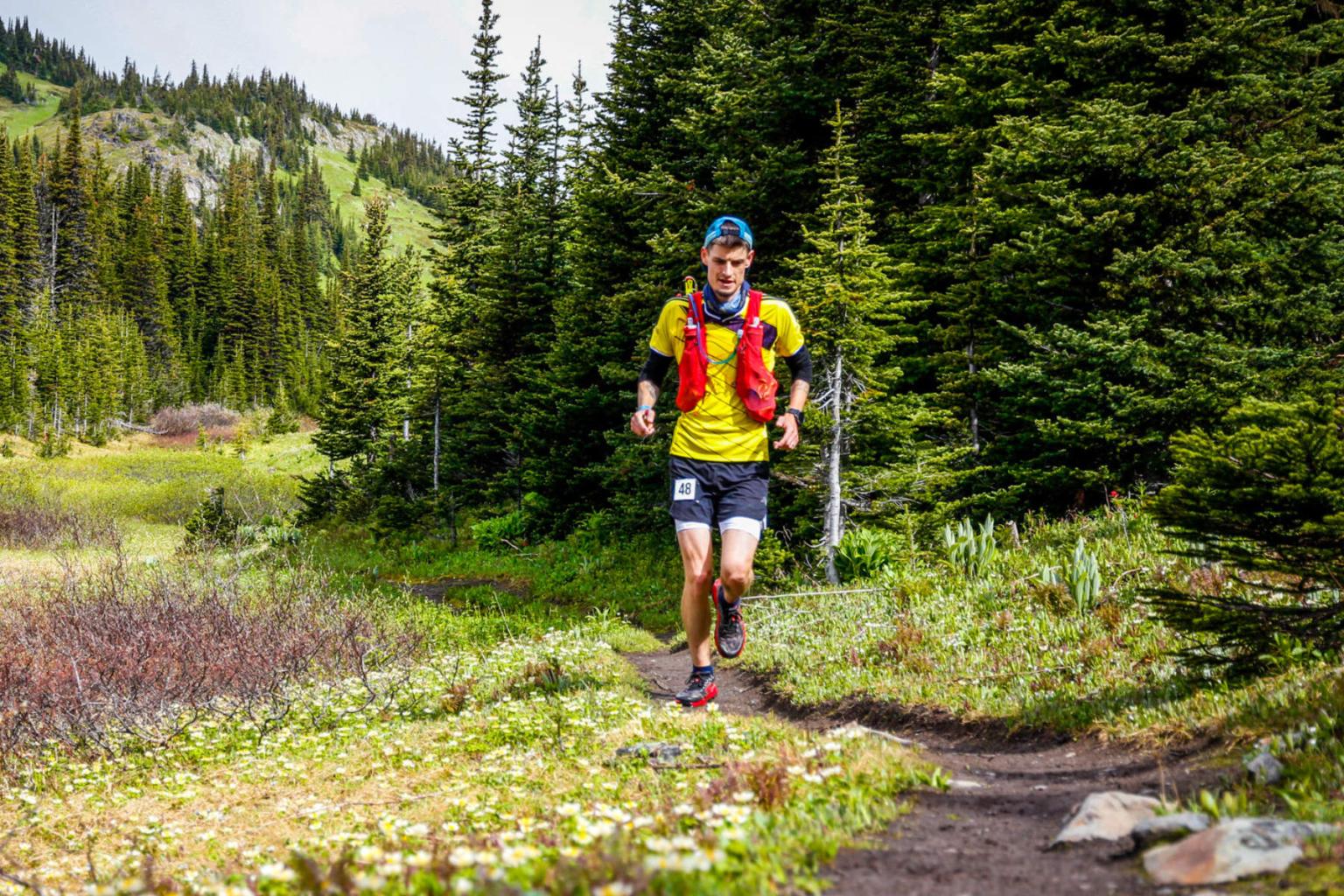 Trail runner in a forest, wearing a red vest and shorts, surrounded by green trees.