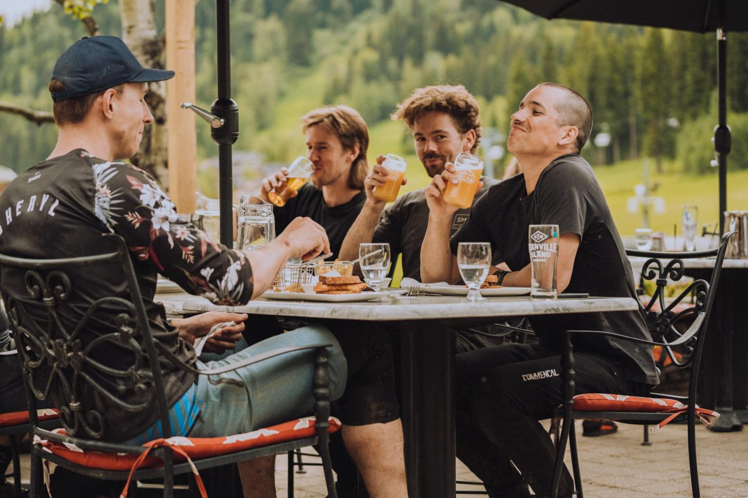 Four men enjoy drinks at an outdoor café with a scenic, green backdrop.