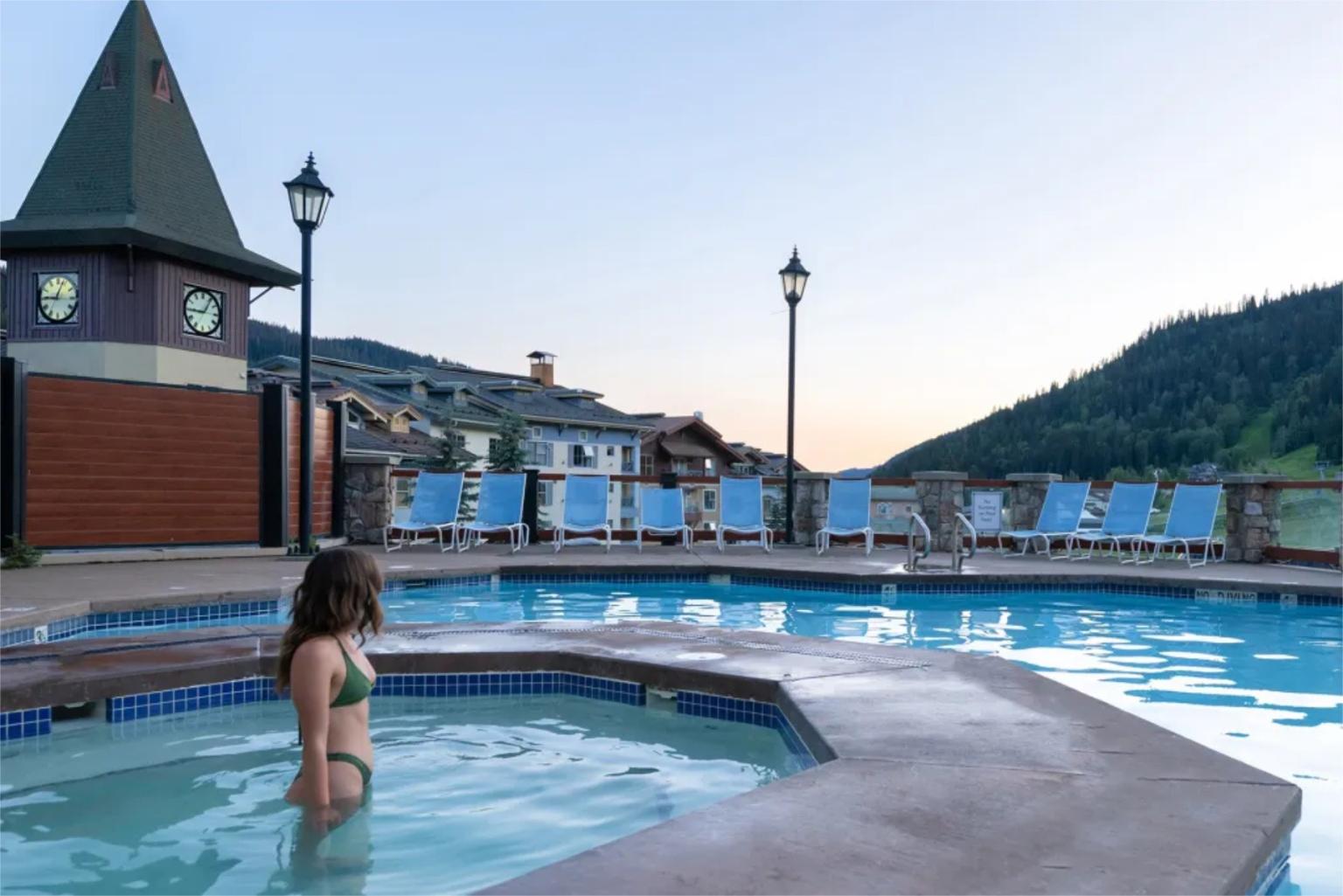 Woman in a hot tub near a pool at sunset, with hills in the background.