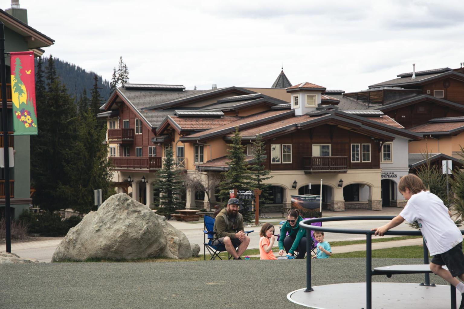 Child playing on merry-go-round, people seated nearby, rustic houses in background.