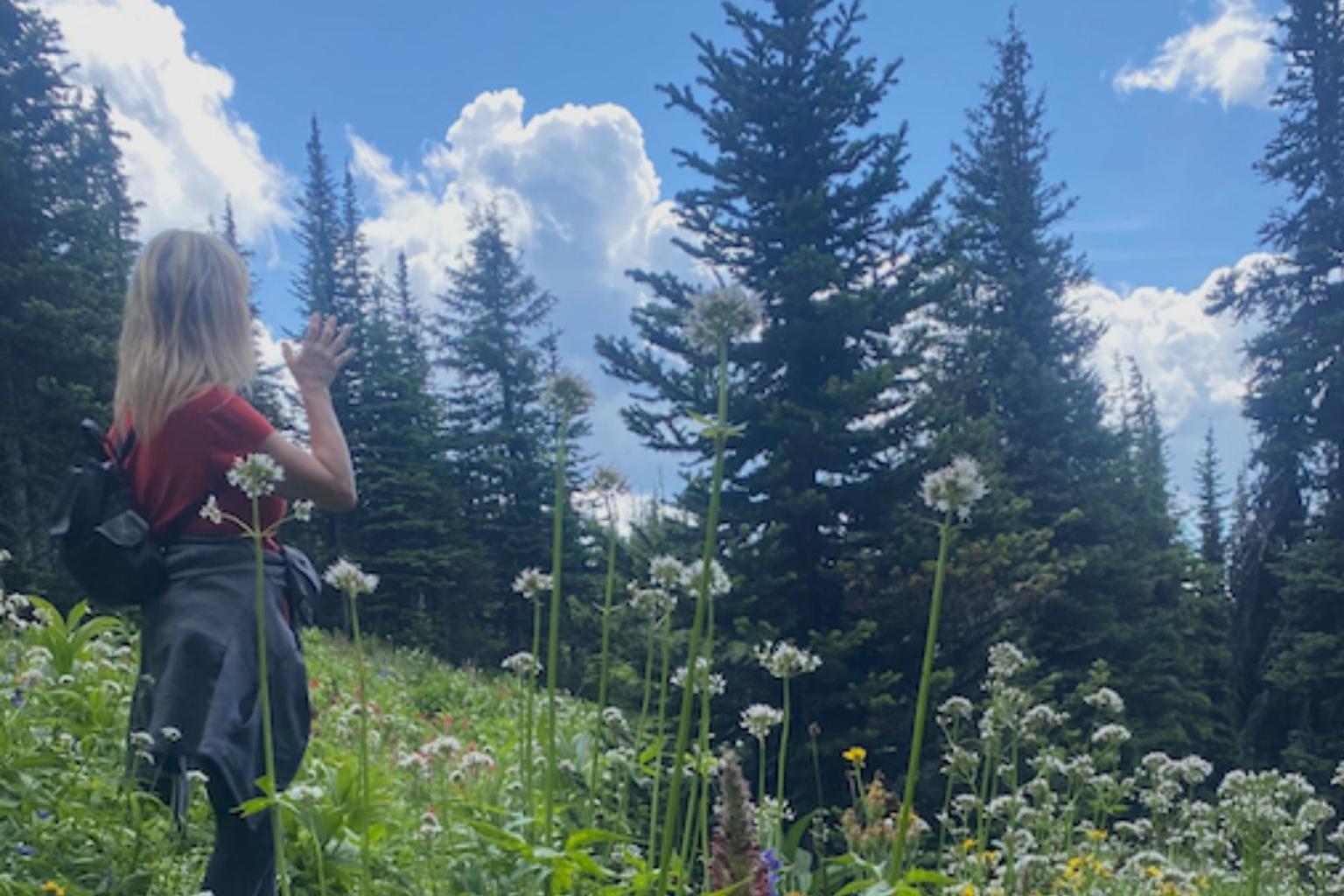 Person in a flower-filled meadow, tall trees, and cloudy blue sky.
