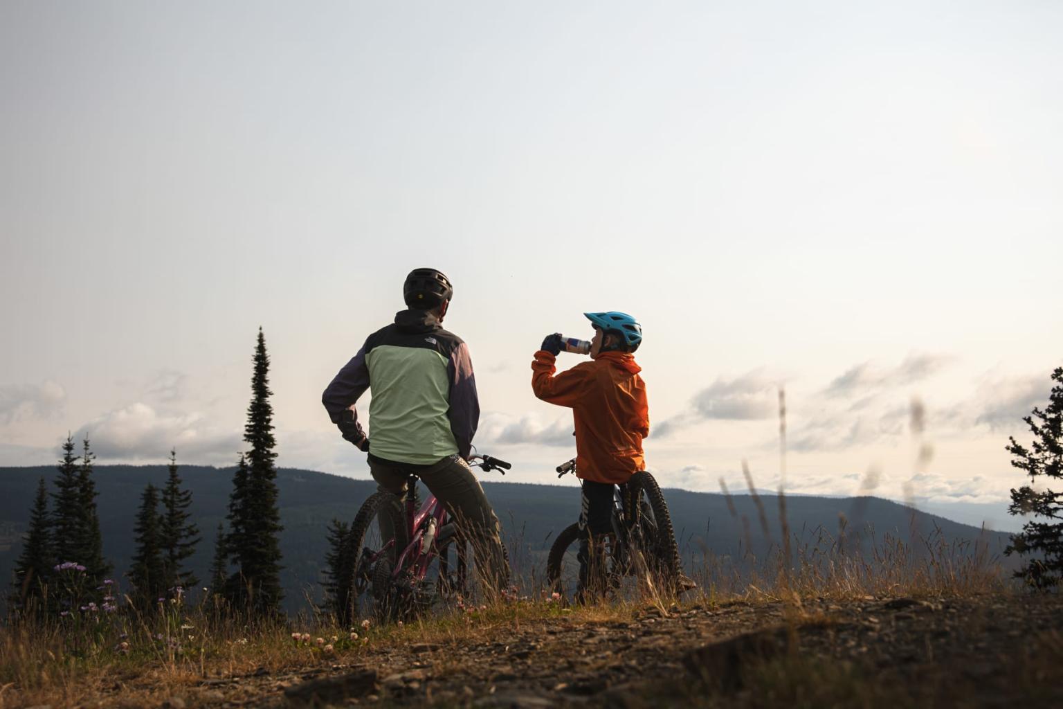 Two cyclists resting on a mountain trail, overlooking a scenic view.