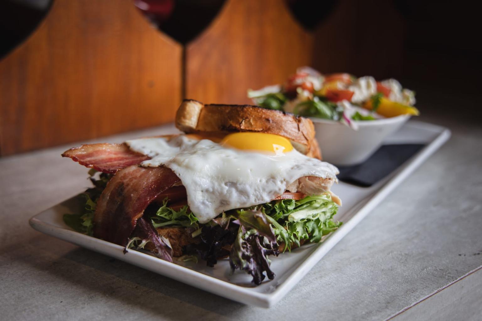 Toasted sandwich with egg, bacon, and greens, with a side salad in soft focus.