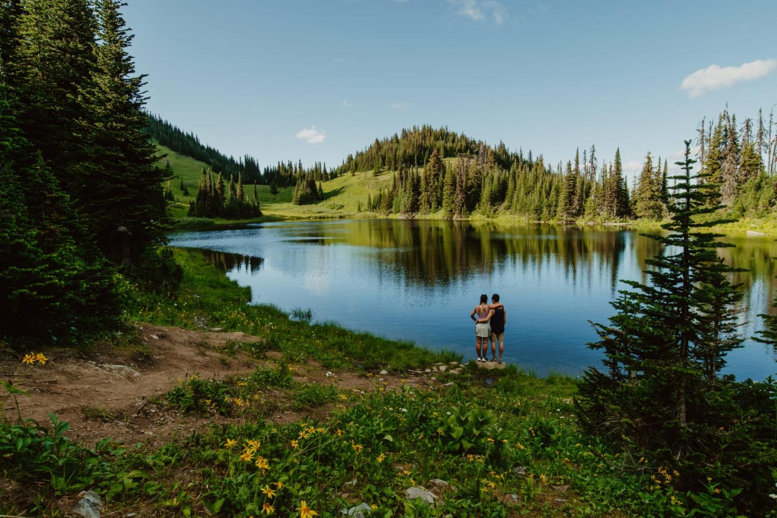 A couple stands by a serene lake surrounded by trees and hills.