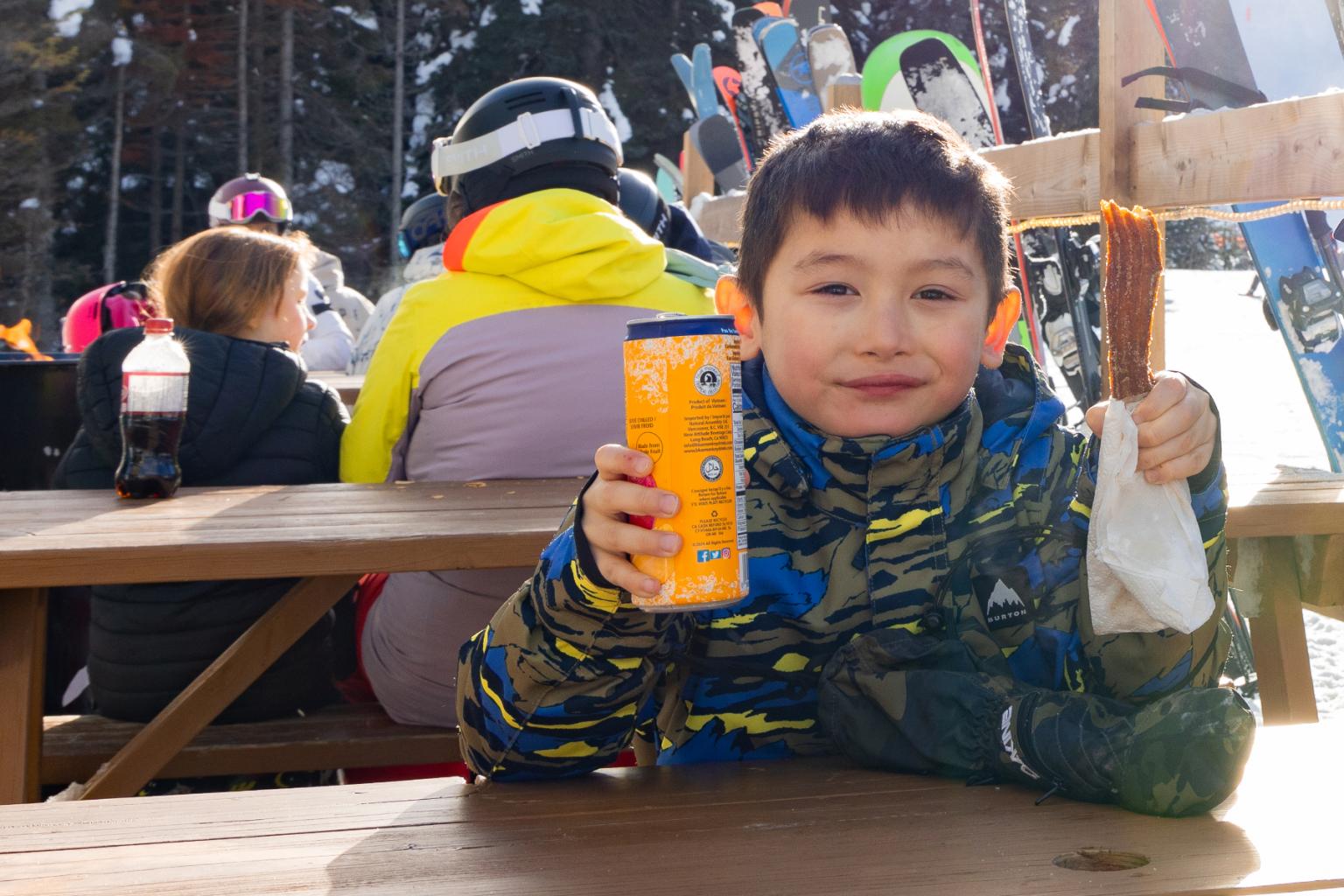 Boy in winter jacket holding a churro and a drink at a ski resort table.
