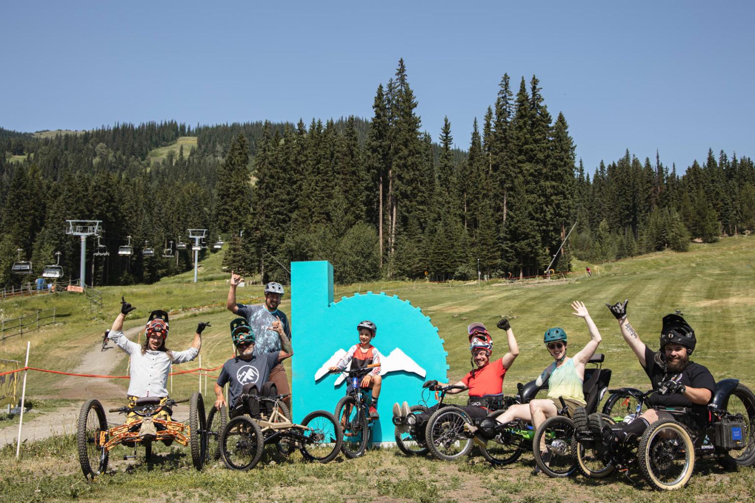 Group in adaptive bikes celebrating outdoors near large turquoise logo.