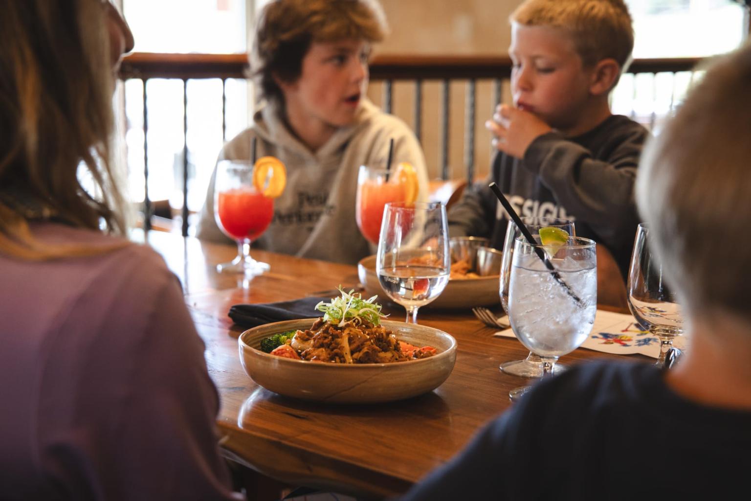 Young people at a wooden table with drinks and pasta dishes.