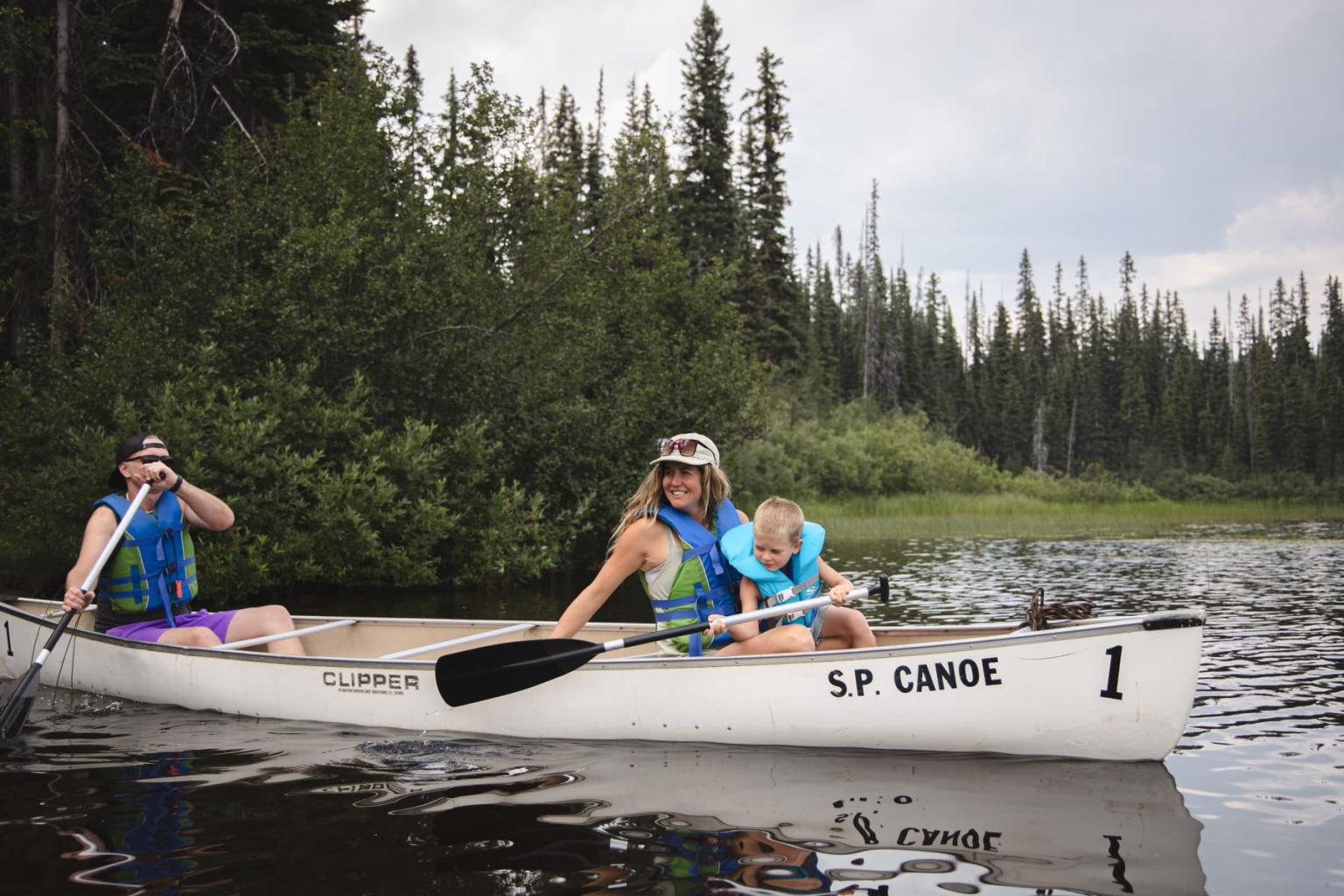 People canoeing on a calm lake surrounded by pine trees.
