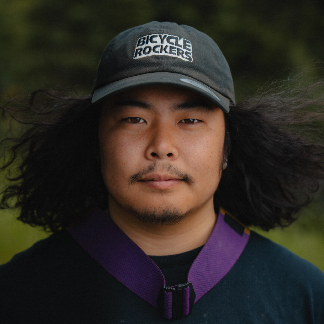 A person with long hair wearing a cap and a purple strap around their neck outdoors.