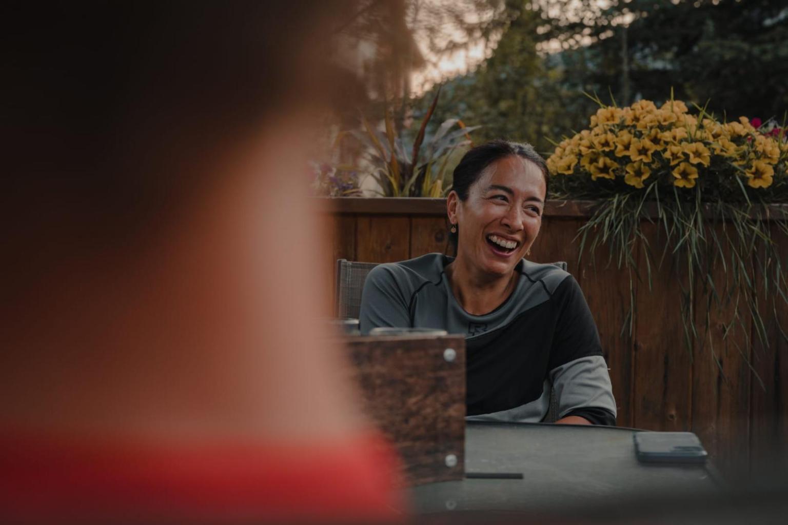 Woman smiling at an outdoor table, yellow flowers in the background.