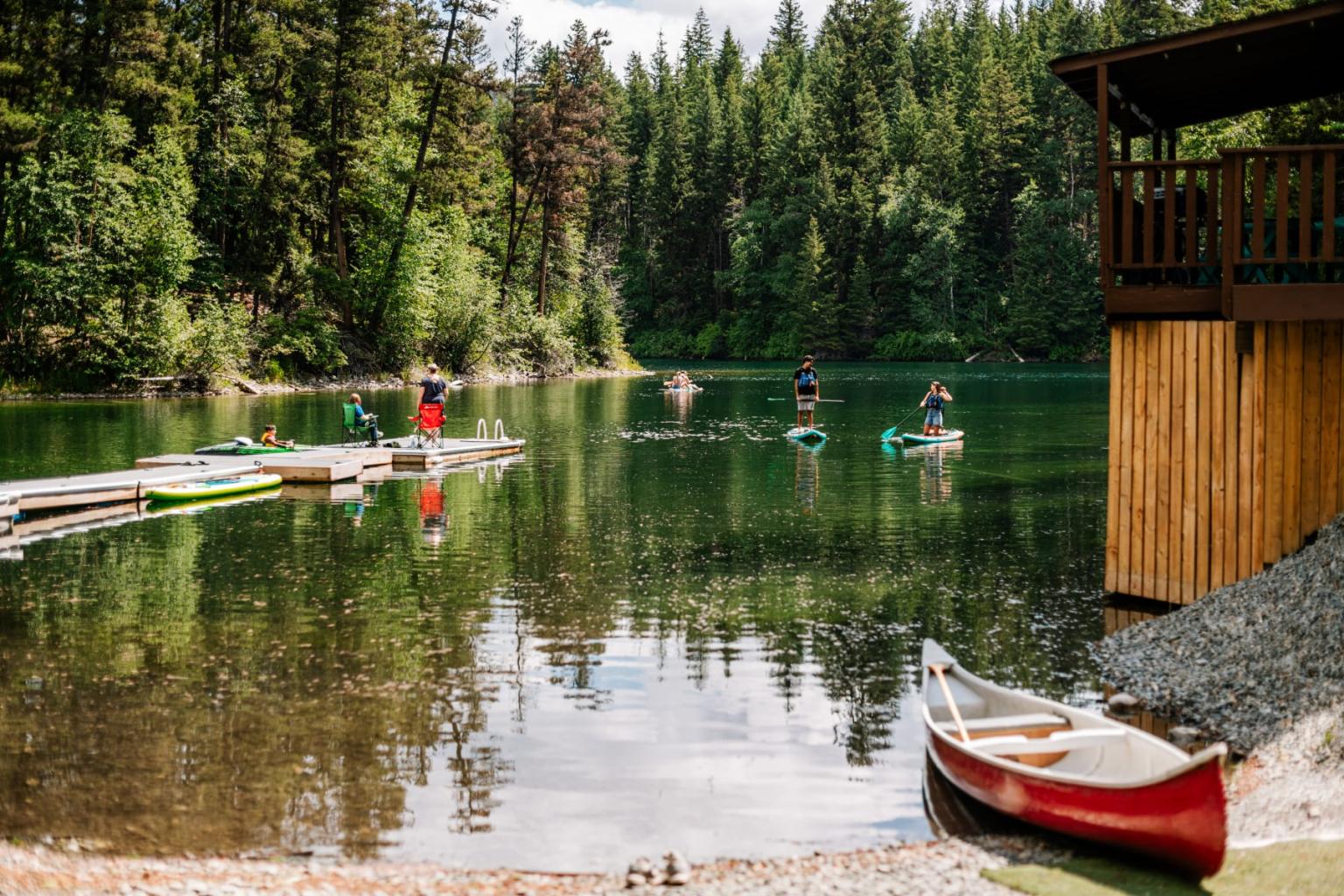 Lake with dock, people paddleboarding, surrounded by trees, red canoe by a wooden building.