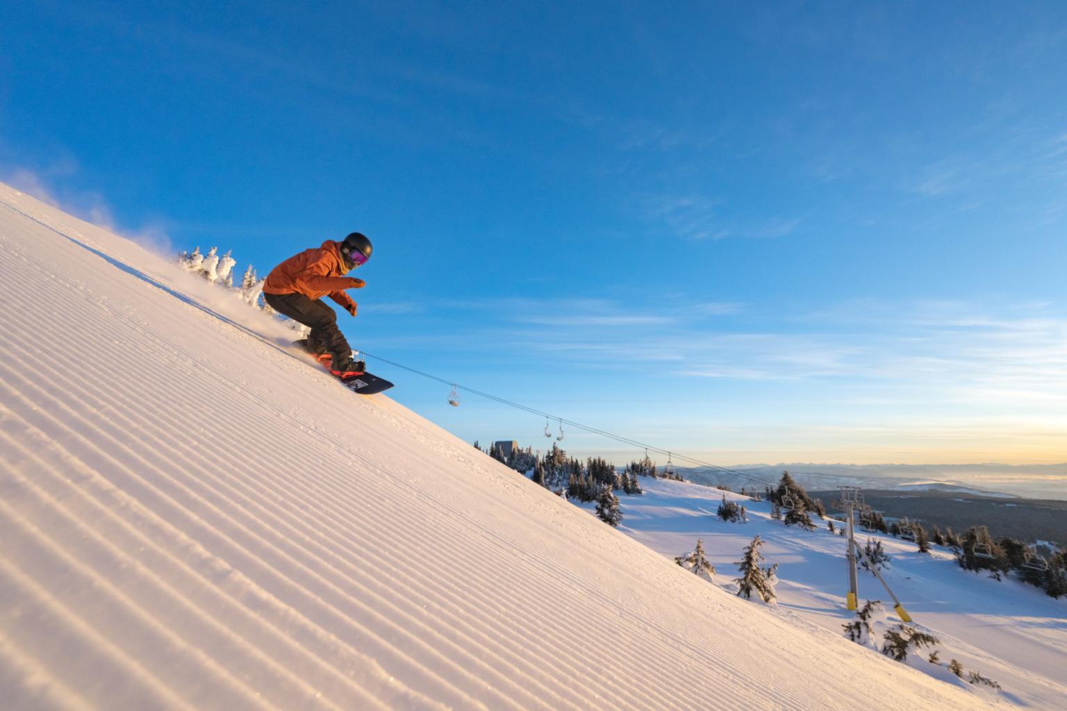 Snowboarder in orange jacket descending a sunny, snow-covered slope.