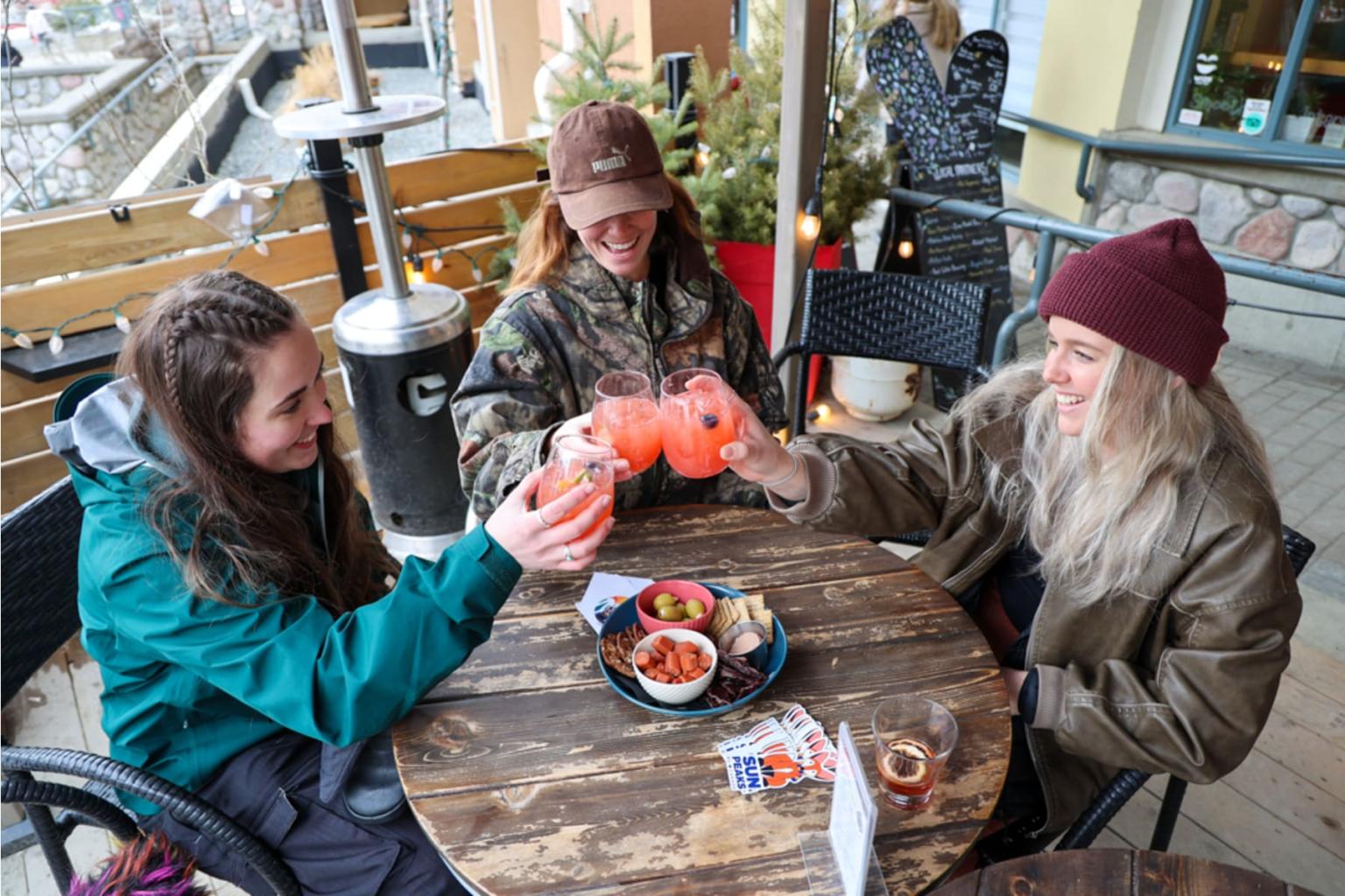 Three people outdoors toast with pink drinks around a wooden table.