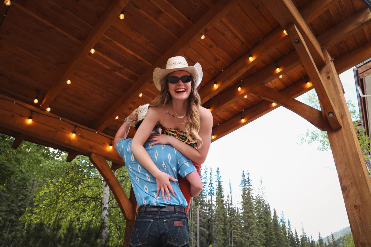 Cowgirl joyfully piggybacks under wooden gazebo with string lights.