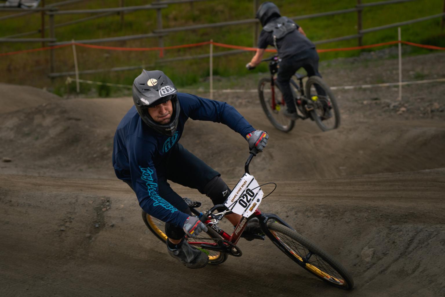 Two cyclists racing on a dirt track, one in foreground and one in background.