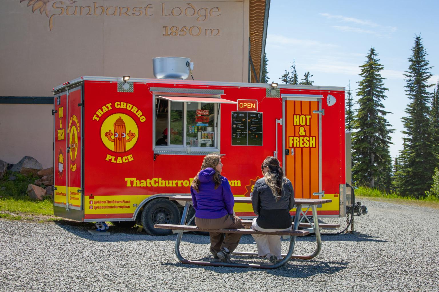 Red food truck with people seated at a picnic table in a sunny outdoor setting.