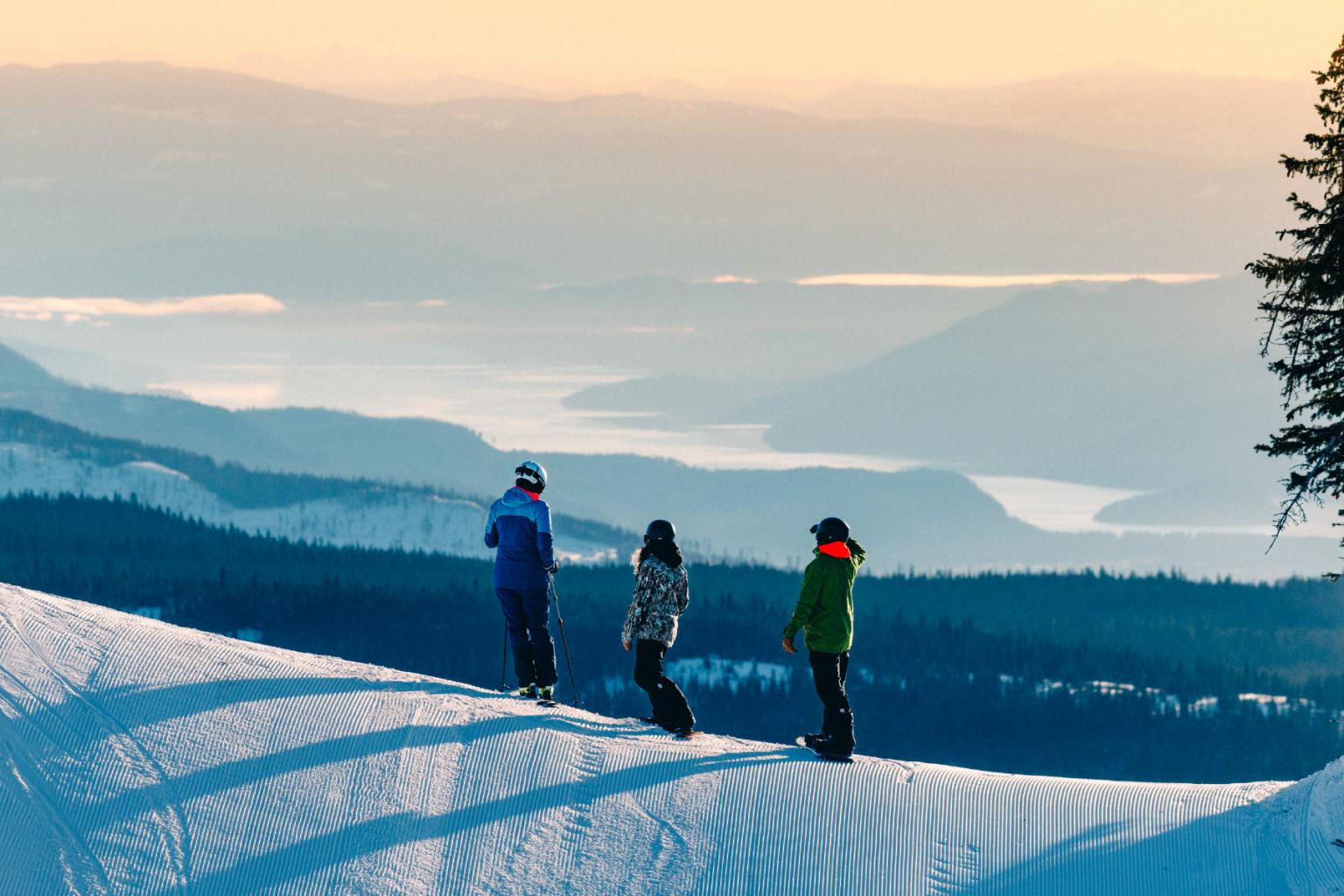 Three people stand on a snowy slope overlooking a scenic mountain view at sunset.
