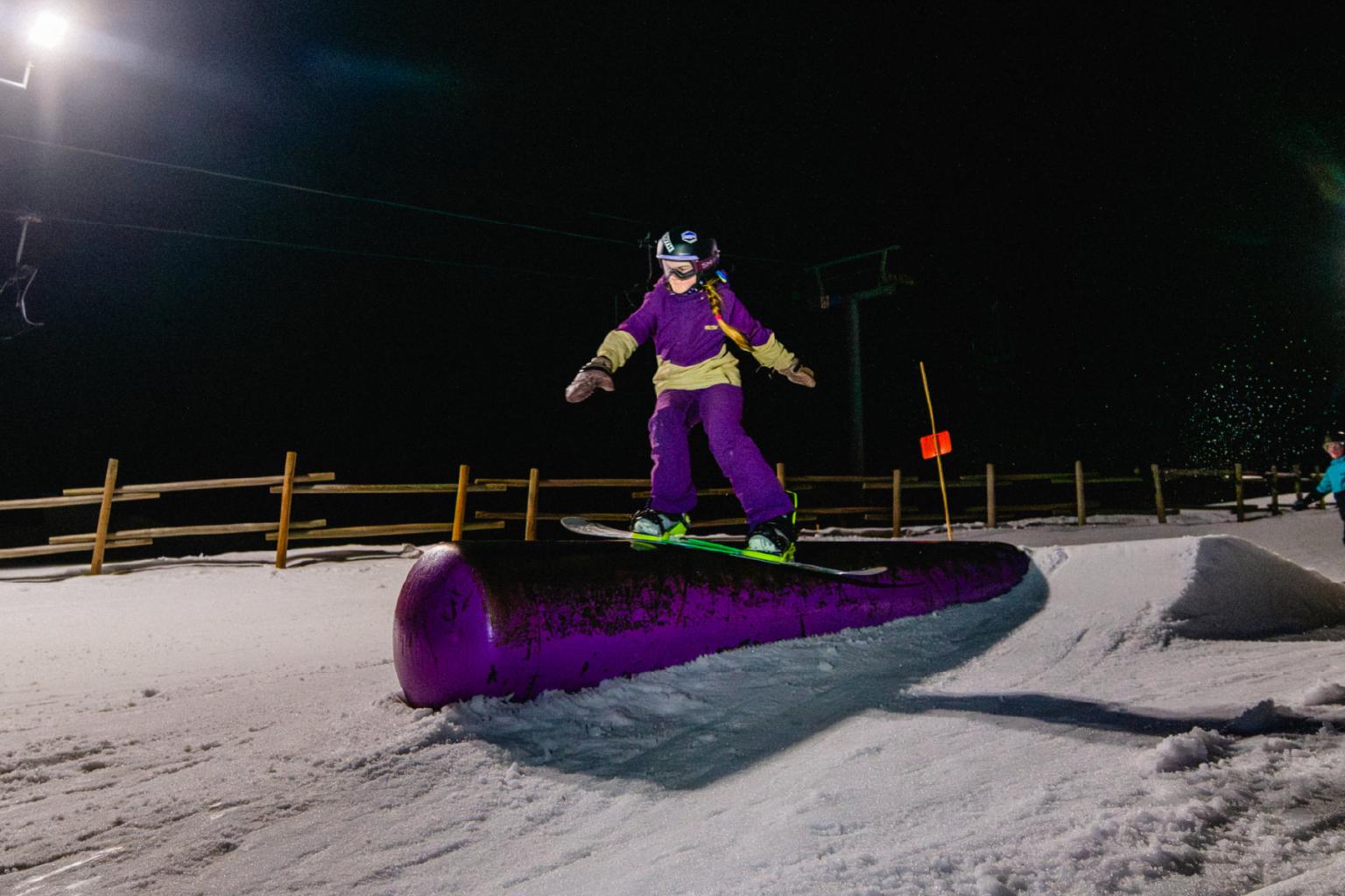 Snowboarder in purple gear gliding over a rail at night on a snowy slope.