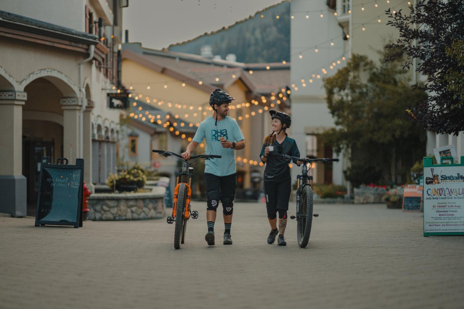 Two people walking bikes under string lights in a quaint town setting.