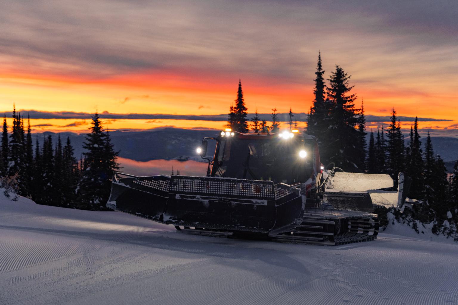 Snow groomer on a snowy mountain at dawn with colorful sky.