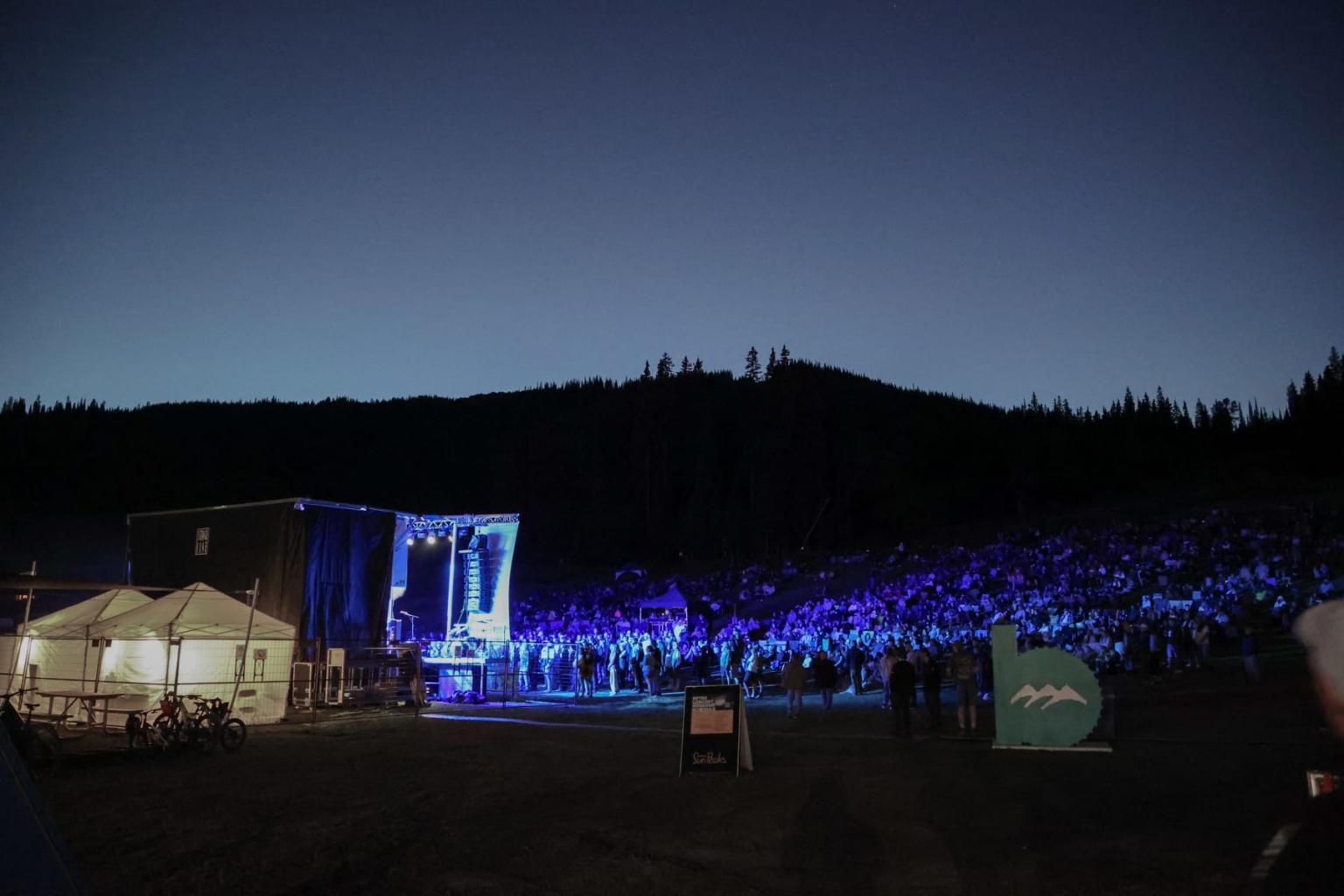 Outdoor concert at dusk, stage lit with blue lights, crowd gathered, mountainous backdrop.