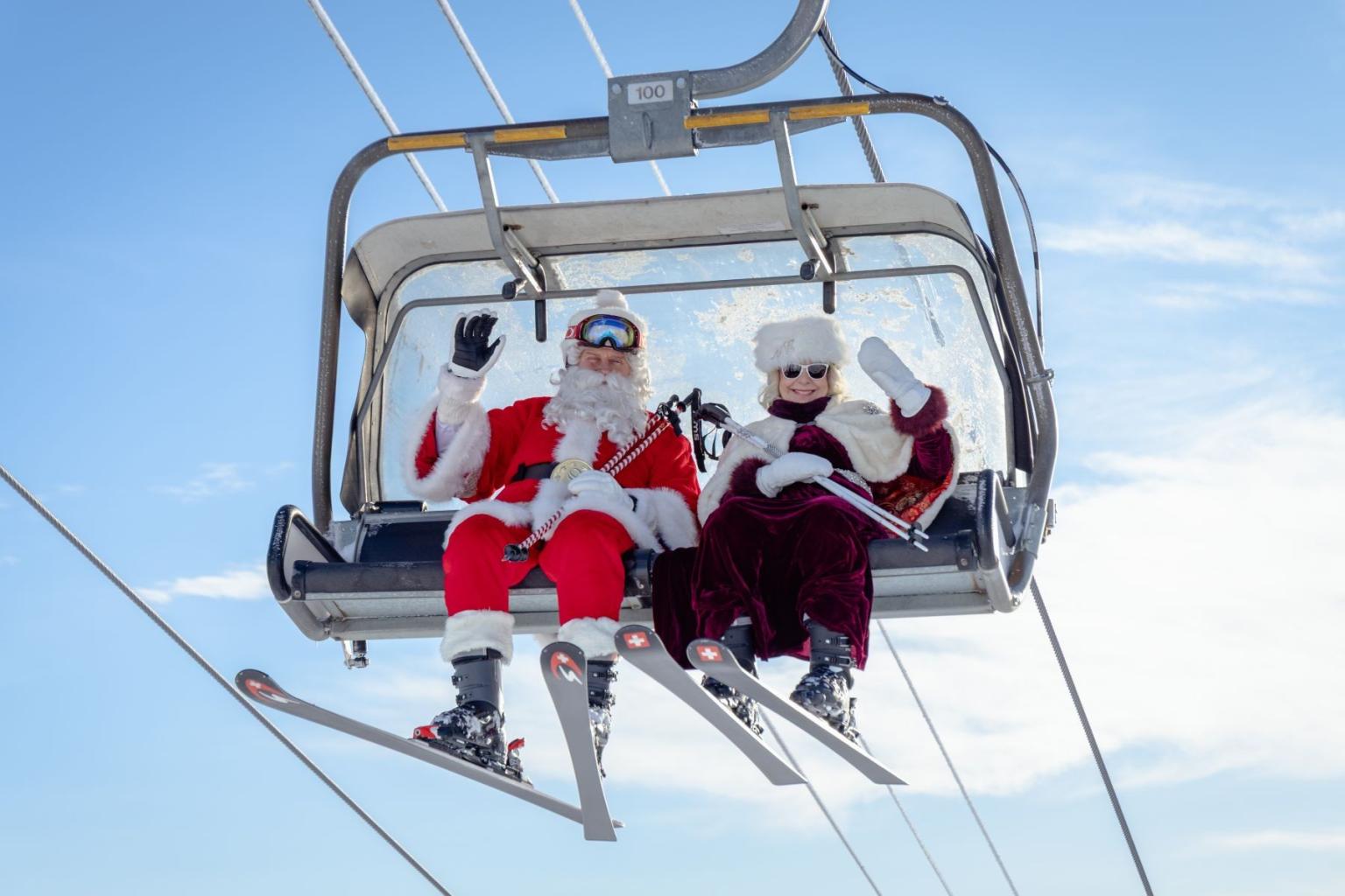 Santa and elf in ski gear on a ski lift, snow-covered mountains behind.