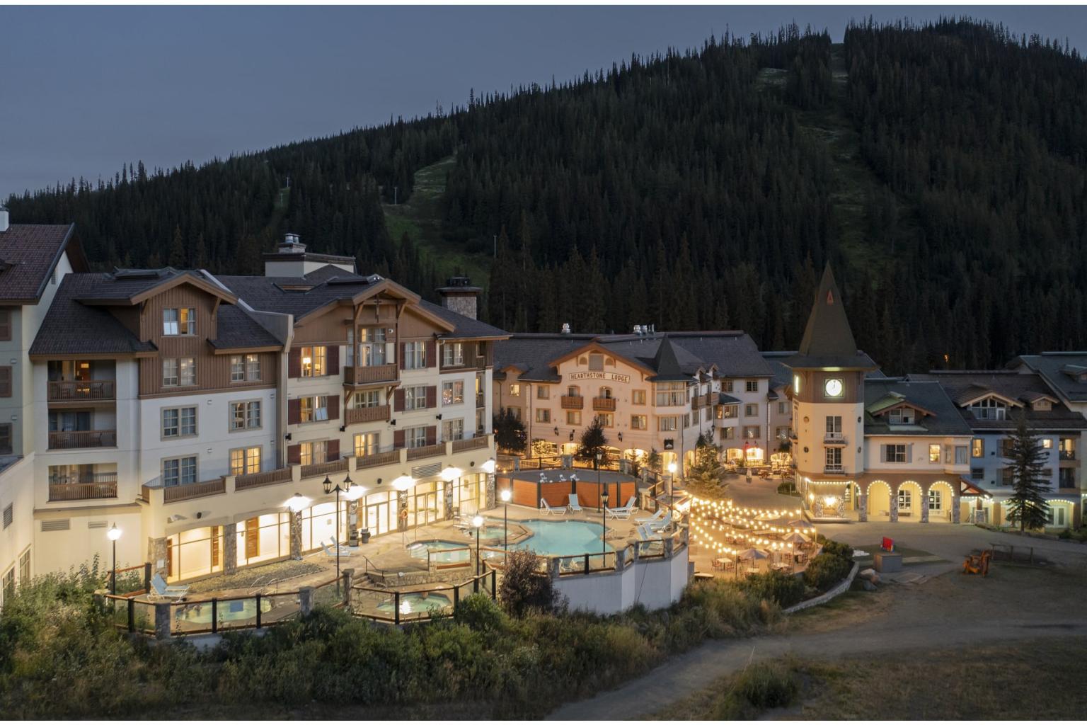 Alpine village at dusk with lit buildings and forested hill backdrop.