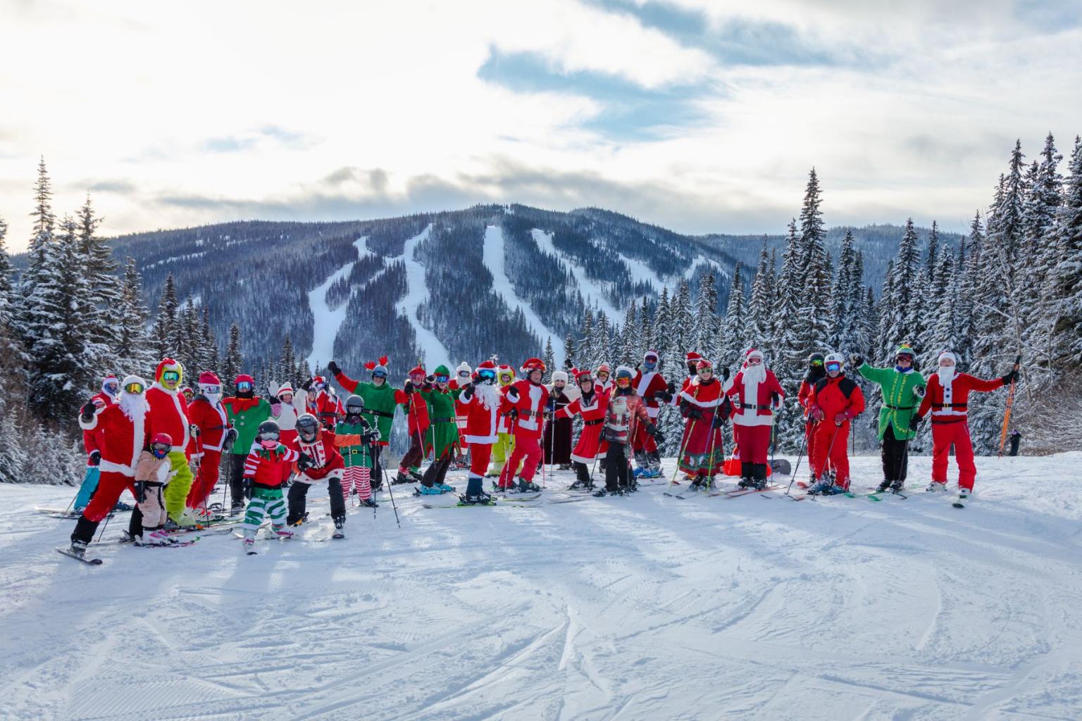 A group dressed in Santa suits pose on a snowy ski slope with mountains in the background.