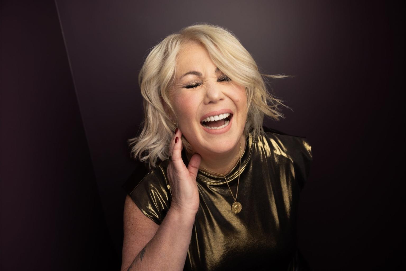 Jann Arden singing in a shiny gold top on a dark background.