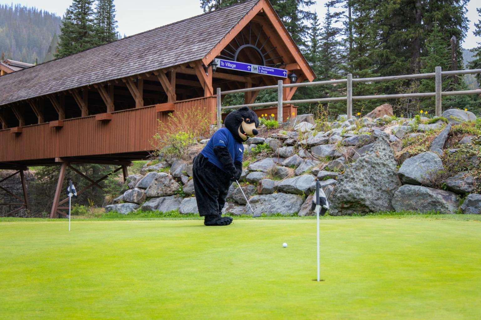 Bear mascot on a golf green beside a rustic wooden bridge.