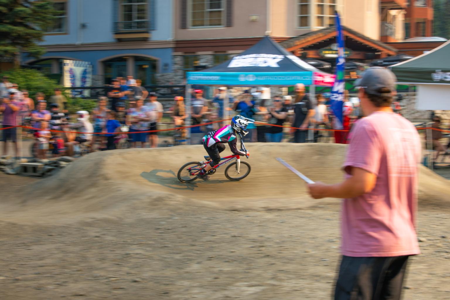 Person racing on a BMX track, watched by spectators and a man in a pink shirt.
