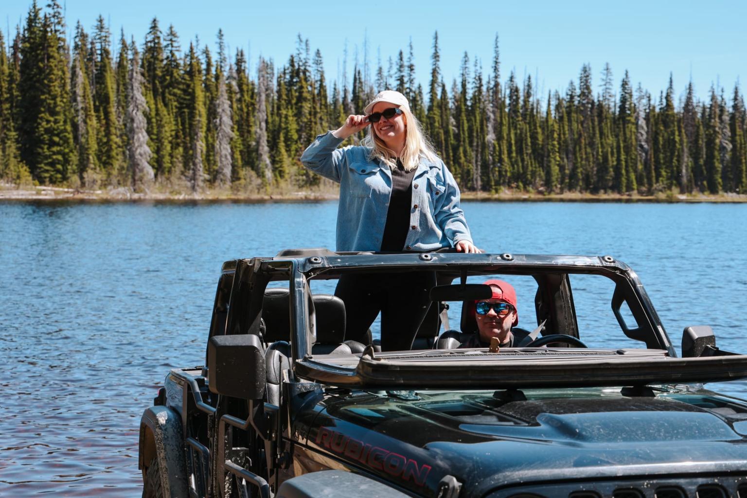 Woman standing in a Jeep by a lake, trees in the background.