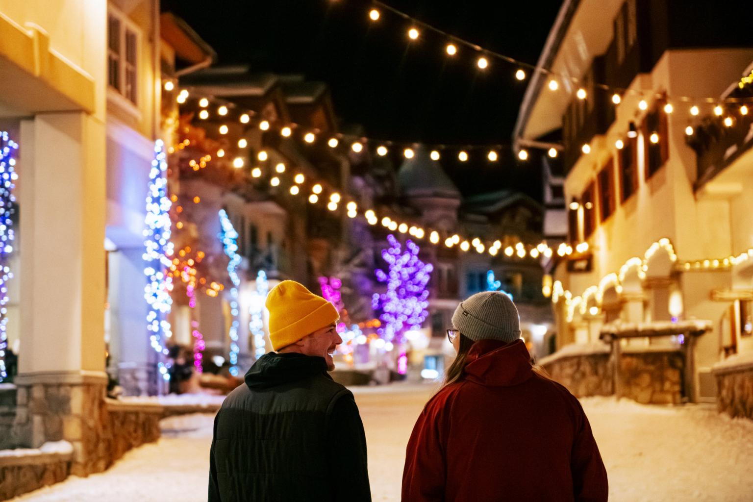 Two people in winter coats walk through a snowy, string-lit village street at night.