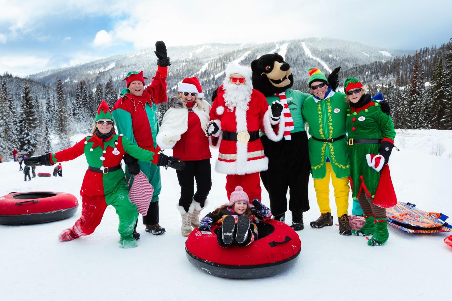 Group in festive costumes posing with snow tubes in a snowy mountain setting.
