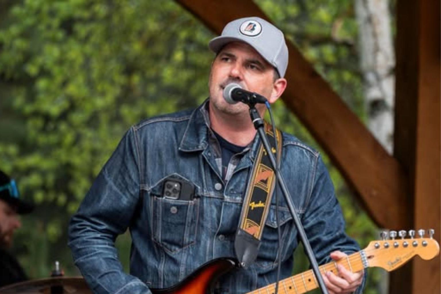 Musician in a cap singing into a microphone while playing electric guitar.