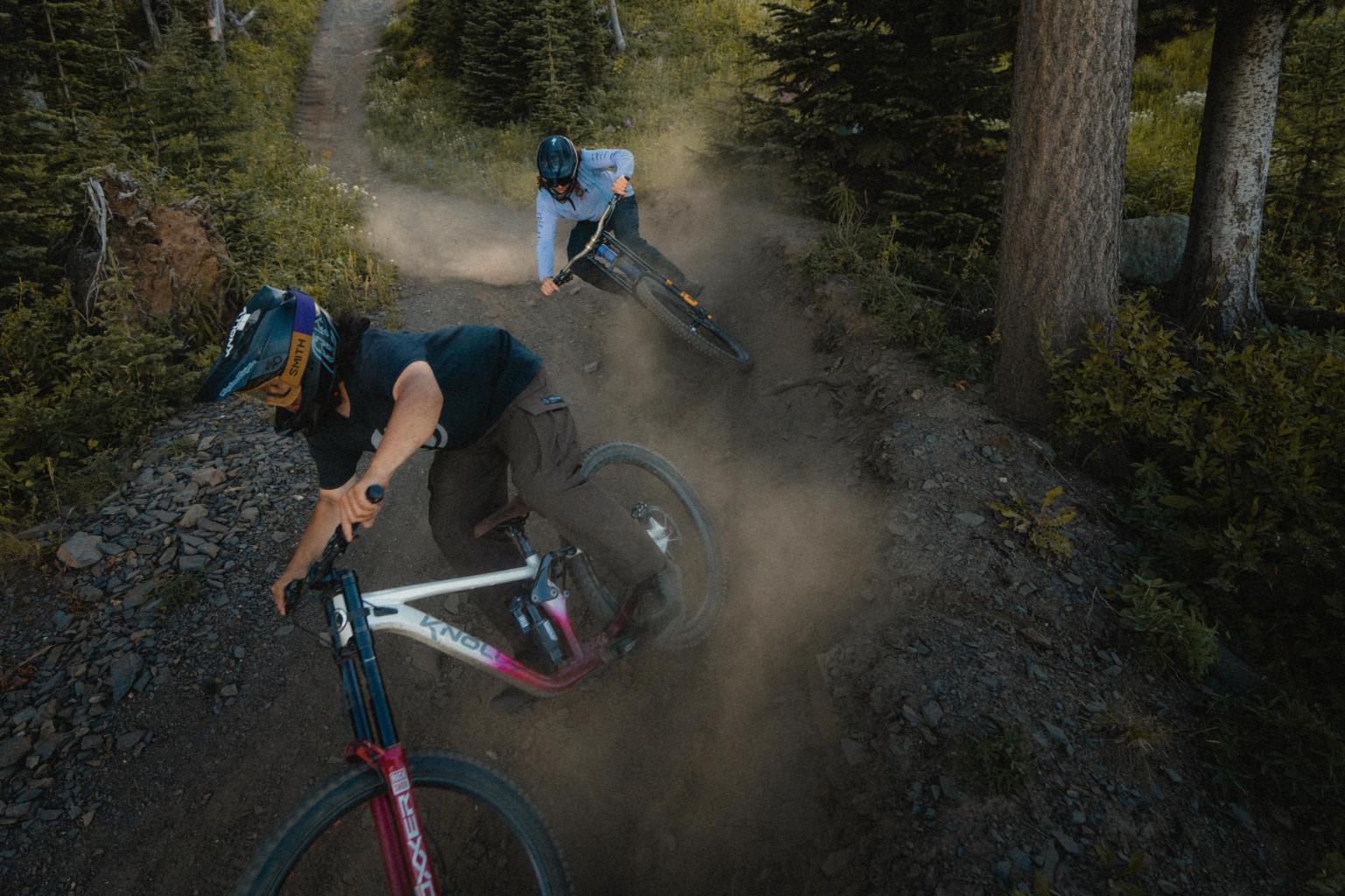 Mountain bikers on a dirt trail through a forest, kicking up dust.