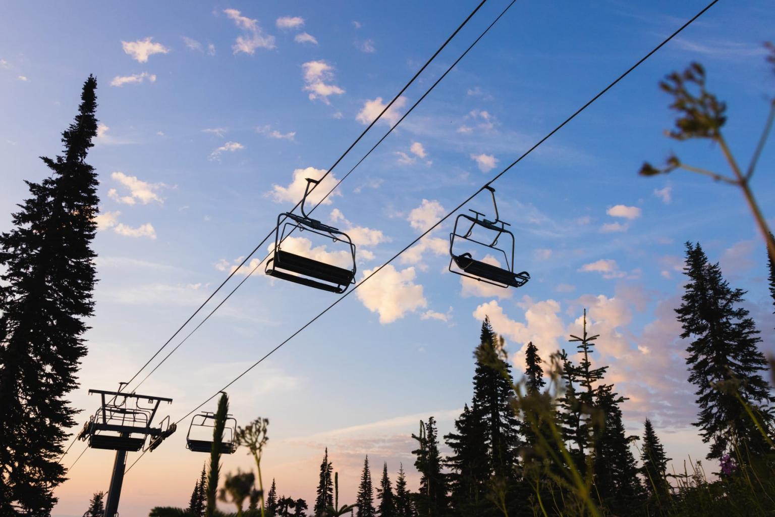 Empty ski lift chairs rise over pine trees at sunset.